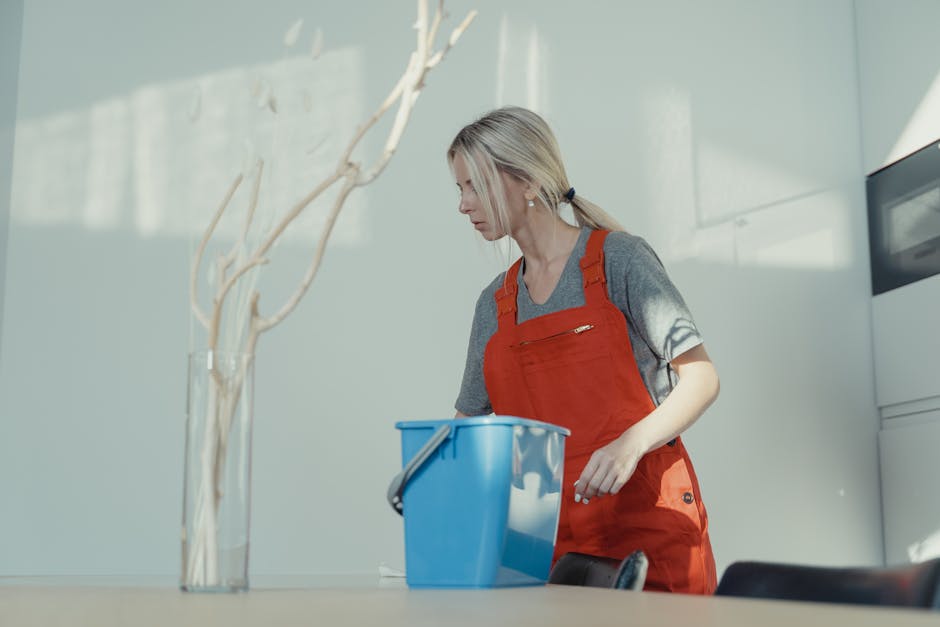 A woman wearing a grey t-shirt and red apron is engaged in surface cleaning inside a modern, well-lit room. She is holding a blue cleaning bucket on a table, which appears to be filled with cleaning supplies. The room features a sleek, minimalist design with a white wall, a tall, white cabinet, and natural daylight streaming through nearby windows, creating a bright atmosphere. The surface she is preparing to clean is a smooth, flat table, likely made of wood or laminate, with a clean and polished finish. A decorative white branch in a clear glass vase is situated on the table, and the overall environment suggests a professional cleaning service setting, emphasizing hygiene, deep cleaning, and surface sanitisation, as provided by Kentish Town Cleaner.