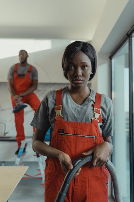 A young woman in a grey t-shirt and red coveralls holding a vacuum cleaner hose, standing in a modern, well-lit room with large windows and a white ceiling. Behind her, a man dressed in similar red workwear is standing on a ladder, possibly engaged in surface cleaning or maintenance work. The room features wooden flooring, a white countertop, and glass surfaces, with natural light illuminating the space. The scene reflects professional domestic or commercial cleaning services provided by Kentish Town Cleaner, emphasizing thorough surface cleaning, hygiene, and deep cleaning processes in a contemporary environment.