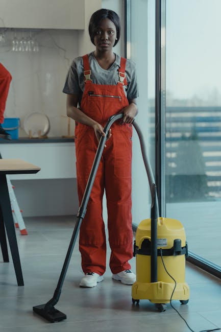 A woman wearing a grey t-shirt and red apron is engaged in surface cleaning inside a modern, well-lit room. She is holding a blue cleaning bucket on a table, which appears to be filled with cleaning supplies. The room features a sleek, minimalist design with a white wall, a tall, white cabinet, and natural daylight streaming through nearby windows, creating a bright atmosphere. The surface she is preparing to clean is a smooth, flat table, likely made of wood or laminate, with a clean and polished finish. A decorative white branch in a clear glass vase is situated on the table, and the overall environment suggests a professional cleaning service setting, emphasizing hygiene, deep cleaning, and surface sanitisation, as provided by Kentish Town Cleaner.