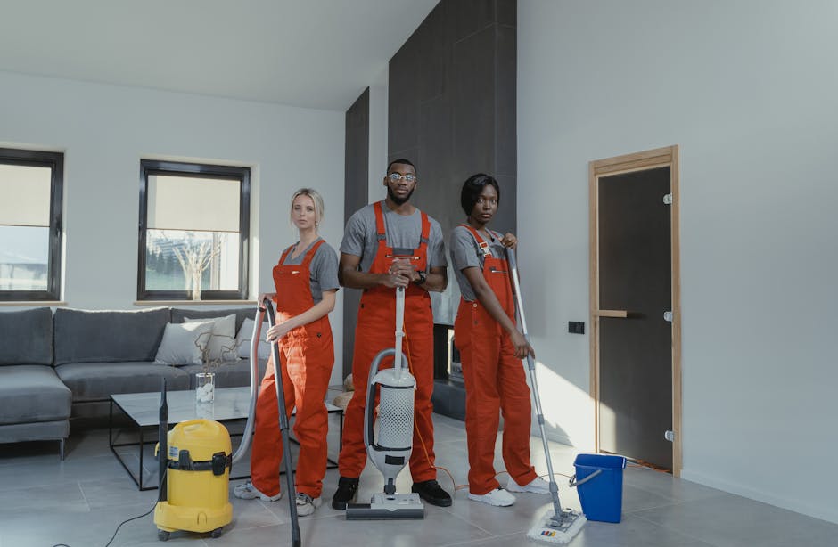 Two professional cleaners wearing orange overalls are performing surface cleaning and deep cleaning in a modern living room, focusing on the light-colored hardwood floor with vacuum and mop tools. The