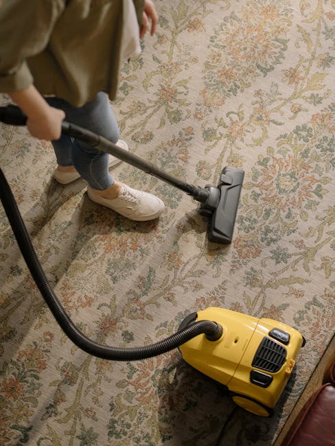 A person wearing beige sneakers and blue jeans is using a vacuum cleaner with a black hose and grey floor head to clean a floral-patterned carpet in a room. The carpet features a design with shades of cream, green, and orange. The room is well lit, showcasing the shiny, clean surface of the carpet. The vacuum cleaner, which appears to be a portable model, is bright yellow with black accents, and the user is holding the hose and extending it across the carpet for thorough surface cleaning. The setting suggests a domestic environment, and the scene highlights ongoing deep cleaning or maintenance as part of regular household hygiene. The background shows a glimpse of furniture, indicating a well-maintained living space, with the presence of natural or artificial lighting enhancing the clarity of the cleaning process. Kentish Town Cleaner specializes in professional domestic cleaning services, including carpet care, as featured on their website ke ntishtowncleaner.co.uk.