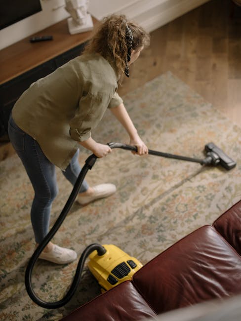 A woman with curly hair, dressed in a green jacket, dark jeans, and white shoes, is engaging in surface cleaning using a yellow vacuum cleaner with a black hose and a wide cleaning nozzle. She is vacuuming a patterned area rug situated in a room with wooden flooring. The lighting is soft and natural, illuminating the beige rug with intricate floral design, and the surrounding space includes a wooden sideboard and a leather sofa in the background. This scene depicts a thorough domestic cleaning process, emphasizing deep cleaning and hygiene as part of the services offered by Kentish Town Cleaner, visible on the webpage for a gig carpet care guide at The Forum Kentish Town.