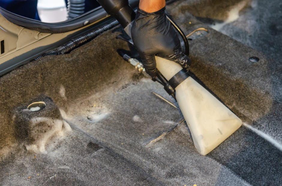 A person wearing beige sneakers and blue jeans is using a vacuum cleaner with a black hose and grey floor head to clean a floral-patterned carpet in a room. The carpet features a design with shades of cream, green, and orange. The room is well lit, showcasing the shiny, clean surface of the carpet. The vacuum cleaner, which appears to be a portable model, is bright yellow with black accents, and the user is holding the hose and extending it across the carpet for thorough surface cleaning. The setting suggests a domestic environment, and the scene highlights ongoing deep cleaning or maintenance as part of regular household hygiene. The background shows a glimpse of furniture, indicating a well-maintained living space, with the presence of natural or artificial lighting enhancing the clarity of the cleaning process. Kentish Town Cleaner specializes in professional domestic cleaning services, including carpet care, as featured on their website ke ntishtowncleaner.co.uk.