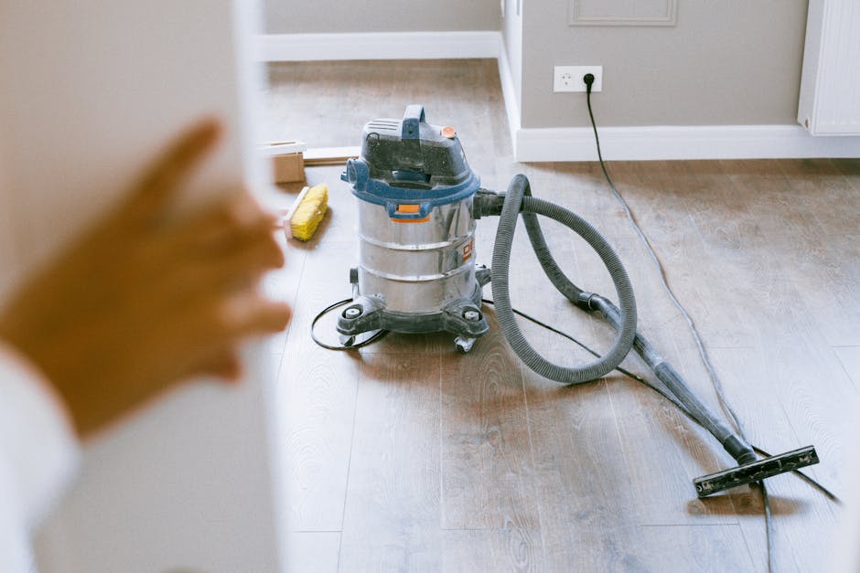 A professional vacuum cleaner with a stainless steel body and black hose attachment is positioned on a wooden floor in a residential room. The room features neutral-colored walls with a white electric
