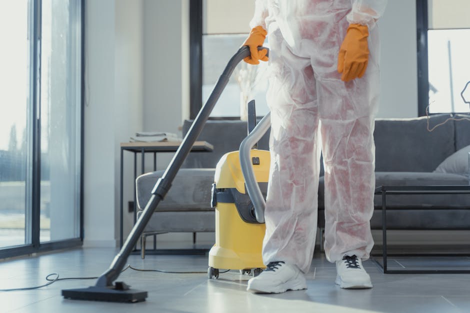 A professional vacuum cleaner with a stainless steel body and black hose attachment is positioned on a wooden floor in a residential room. The room features neutral-colored walls with a white electric